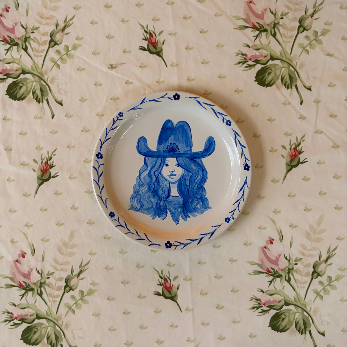 A small round ceramic plate with a blue illustration of a woman cowgirl in a cowboy hat, placed on a floral tablecloth with pink roses and green leaves.