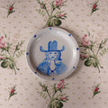 A white ceramic plate featuring a blue painted illustration of a woman in a cowboy hat, surrounded by small blue stars, displayed on a beige floral tablecloth.
