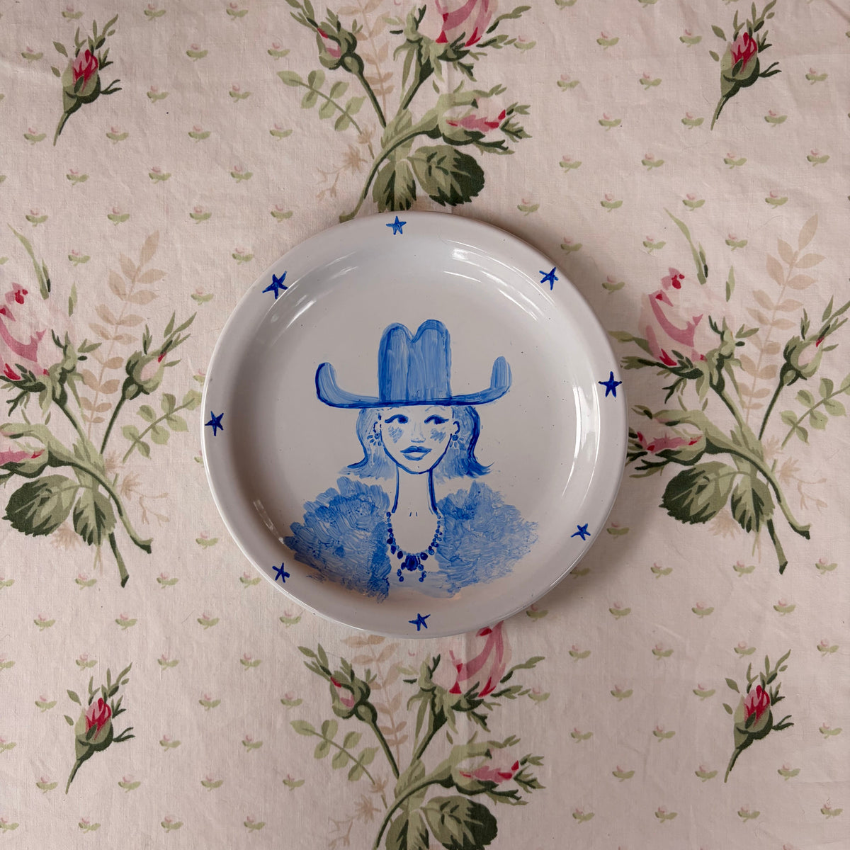 A white ceramic plate featuring a blue painted illustration of a woman in a cowboy hat, surrounded by small blue stars, displayed on a beige floral tablecloth.