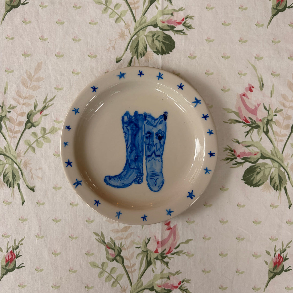 A small round ceramic plate with a blue boot illustration and blue stars around the rim, placed on a floral tablecloth with pink roses and green leaves.