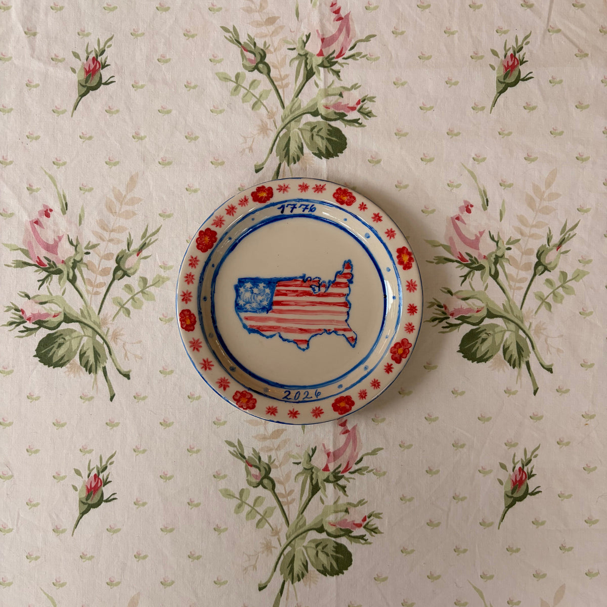 hand painted map of America with American flag 1776 2026 with groovy red and flower with orange center border on small ivory plate with colors red blue and orange