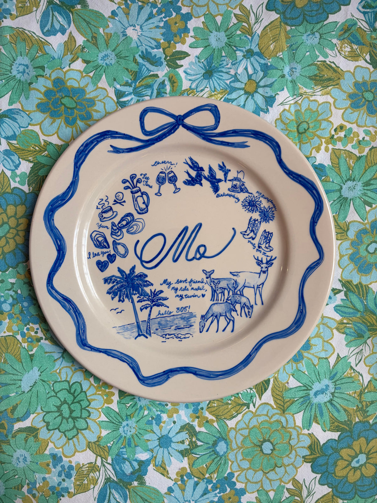 A round ceramic plate with a scalloped rim, featuring blue illustrations of favorite things and the cursive name 'Mo' in the center, laid flat on a green and blue floral patterned fabric background.
