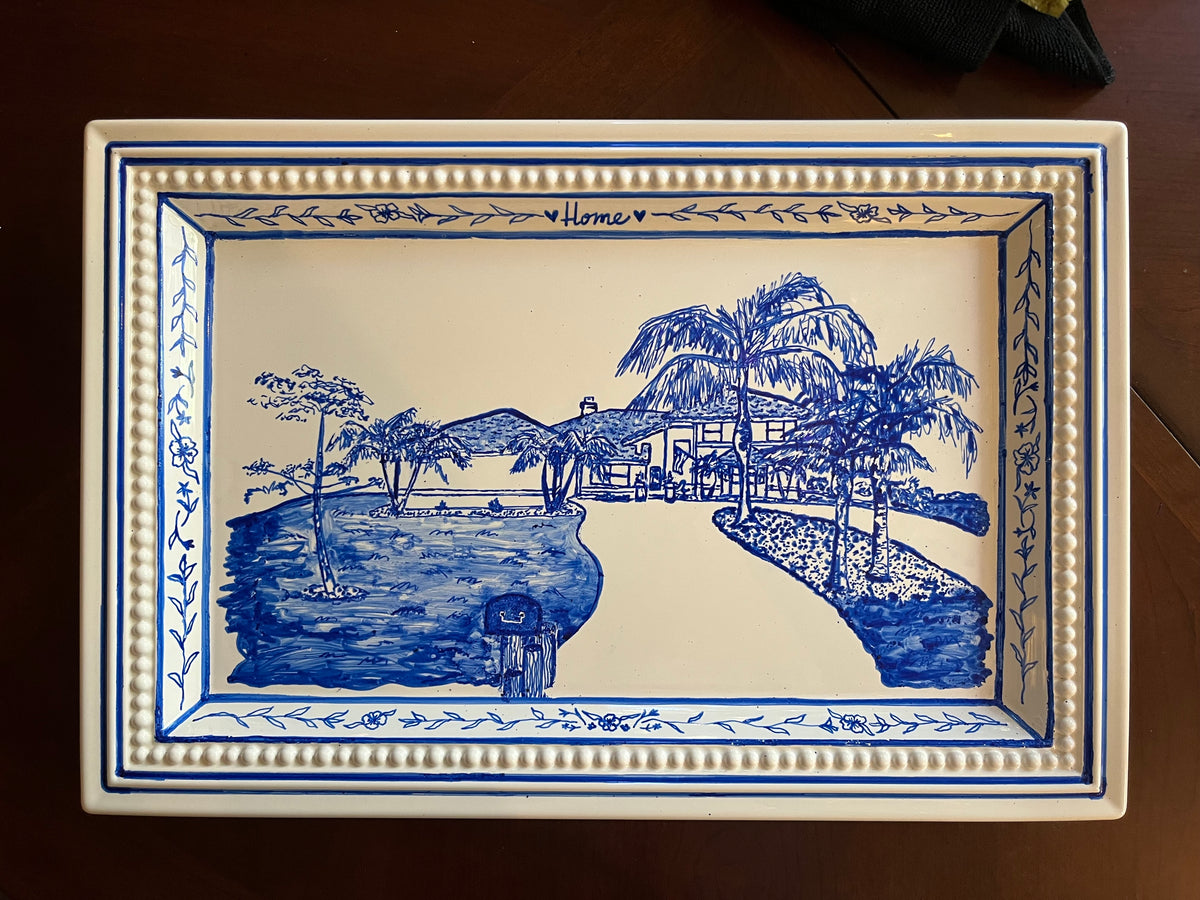 A rectangular ceramic serving tray with a blue and white landscape illustration and the word 'Home' written at the top, placed on a dark wooden surface.