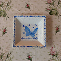 A square white ceramic dish with a blue butterfly illustration and floral border, placed on a beige floral tablecloth.
