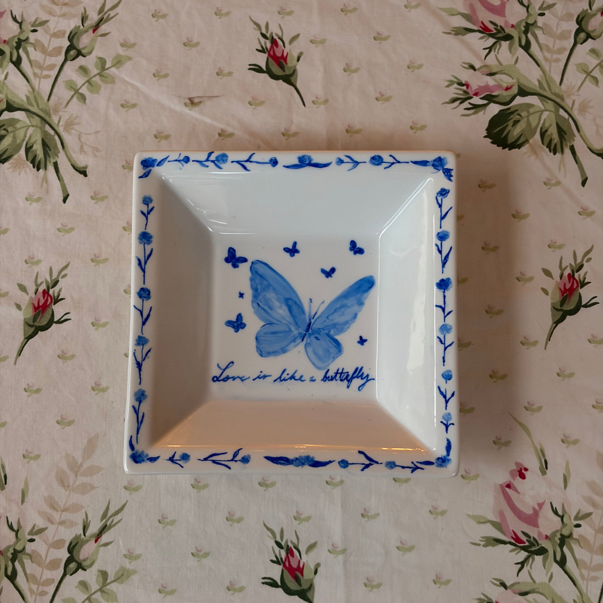 A square white ceramic dish with a blue butterfly illustration and floral border, placed on a beige floral tablecloth.