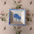 A square white ceramic dish with a blue palm tree design in the center and a blue striped rim, placed on a beige floral tablecloth.