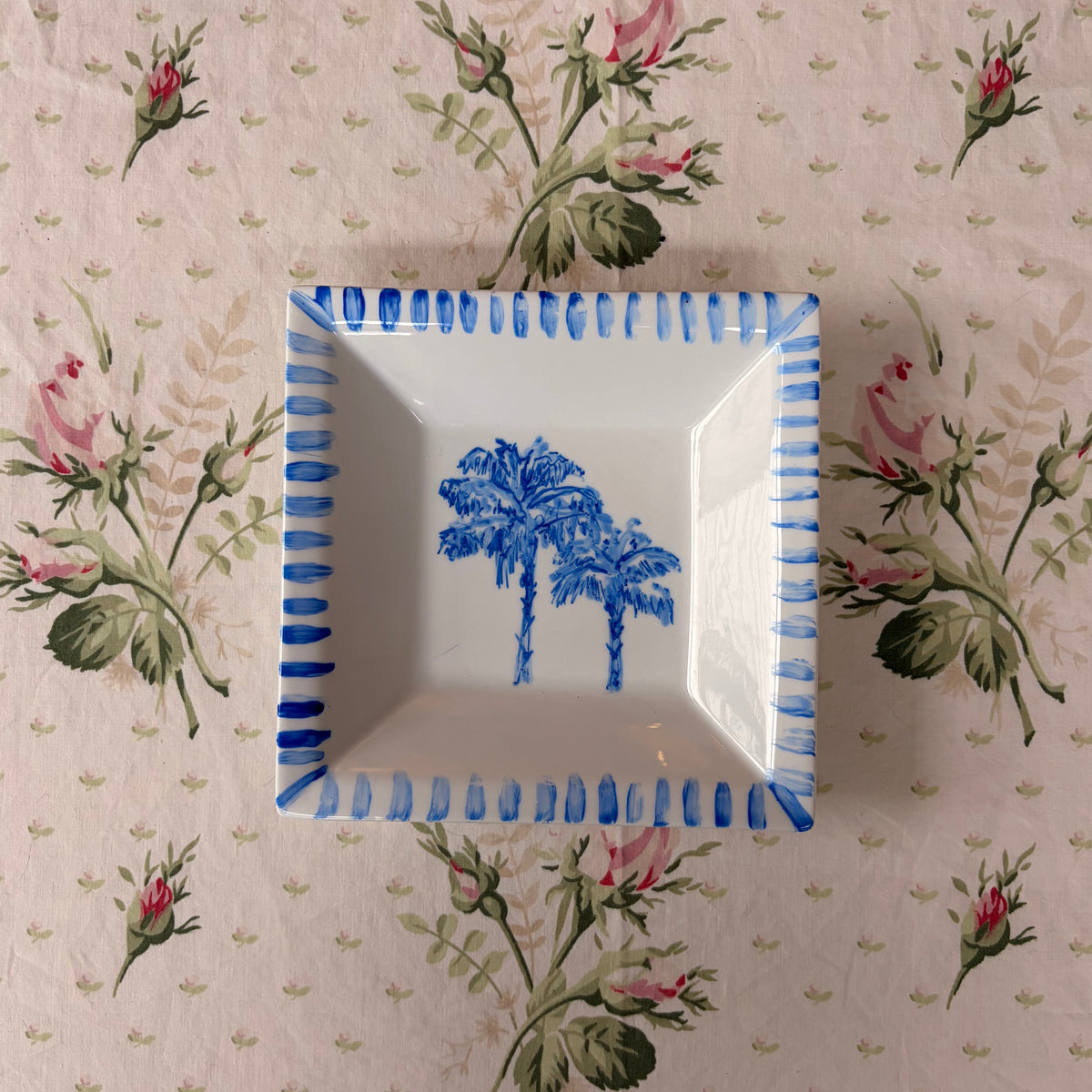 A square white ceramic dish with a blue palm tree design in the center and a blue striped rim, placed on a beige floral tablecloth.