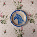 A round ceramic plate with a blue scalloped rim and a painted blue horse head, placed on a pink and green floral tablecloth.