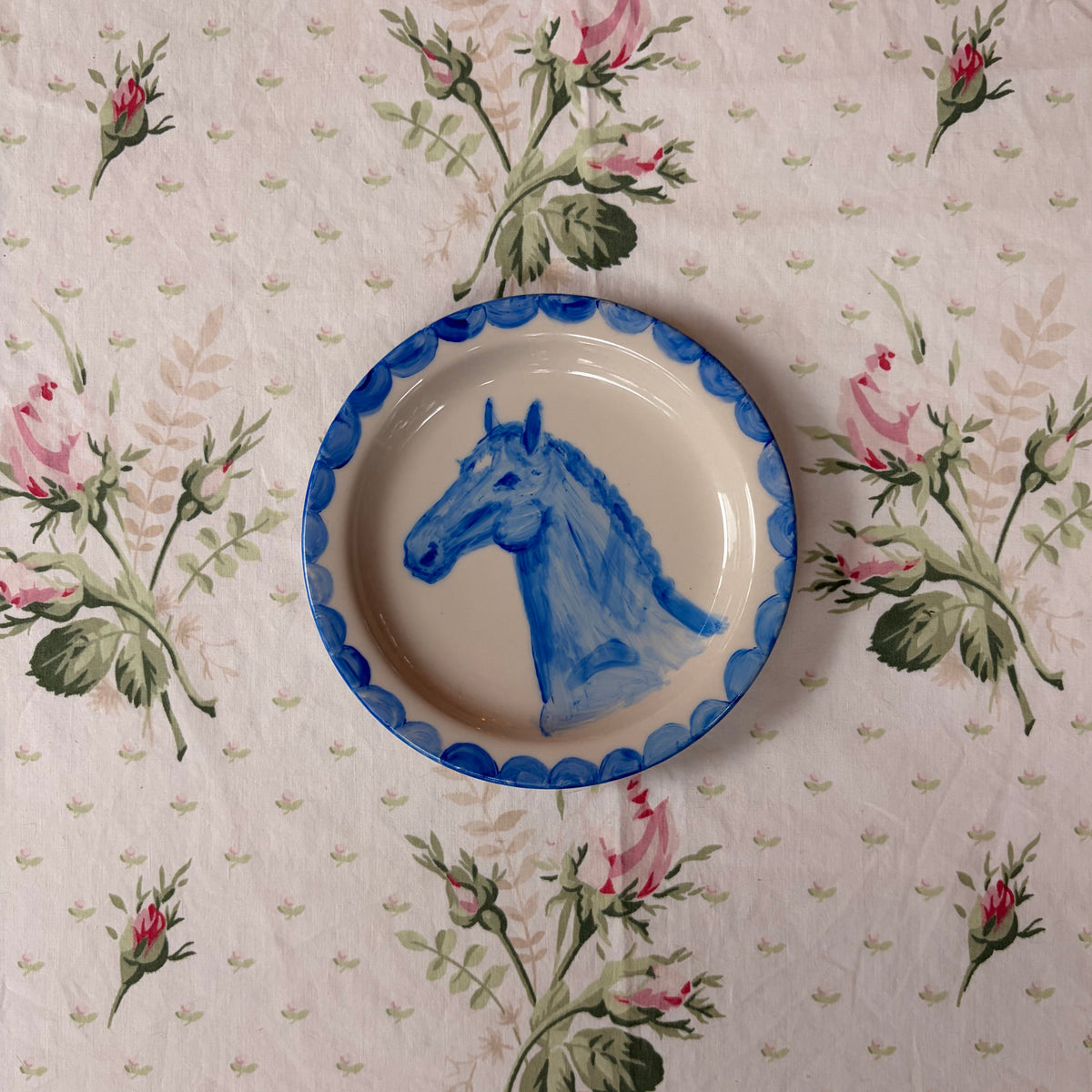 A round ceramic plate with a blue scalloped rim and a painted blue horse head, placed on a pink and green floral tablecloth.