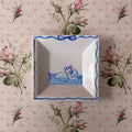 A square white ceramic dish with blue decorative borders and a swan illustration, placed on a pink floral tablecloth.