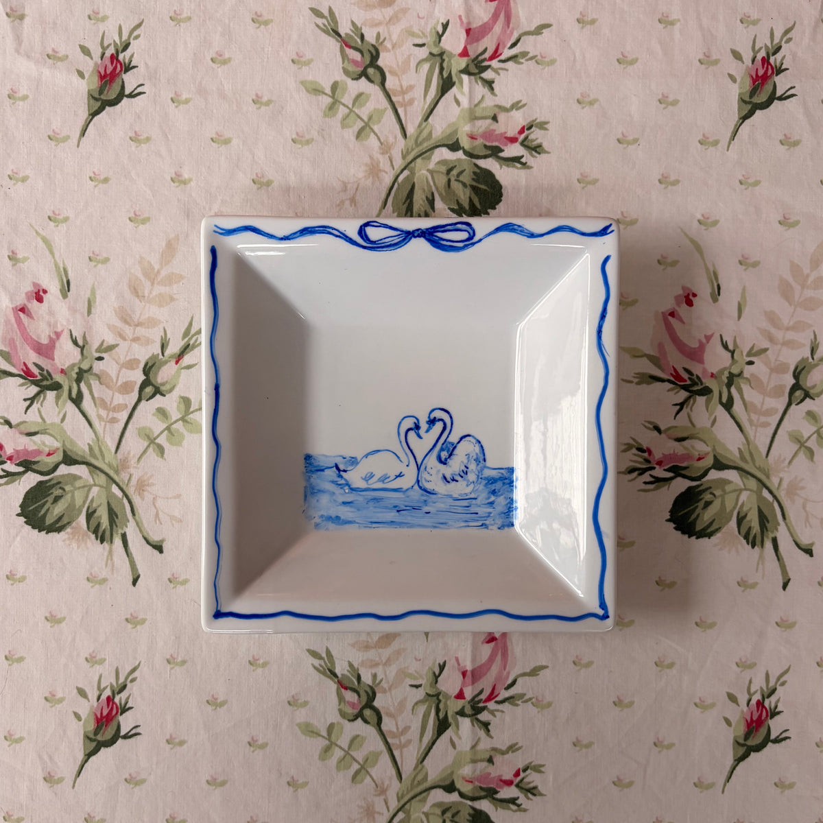 A square white ceramic dish with blue decorative borders and a swan illustration, placed on a pink floral tablecloth.