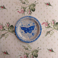 A round ceramic plate with a blue butterfly design and blue scalloped rim, placed on a floral tablecloth with pink roses and green leaves.