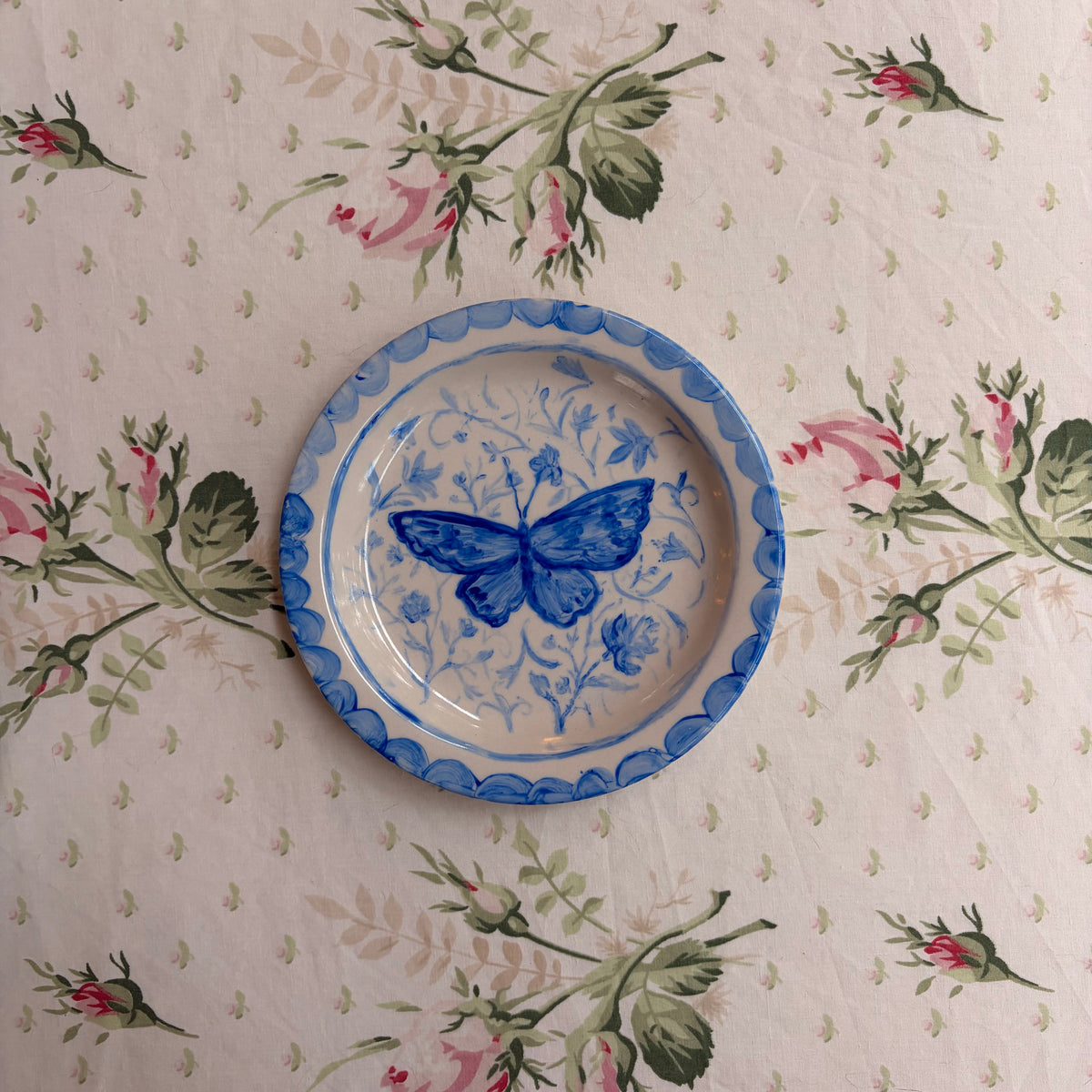 A round ceramic plate with a blue butterfly design and blue scalloped rim, placed on a floral tablecloth with pink roses and green leaves.