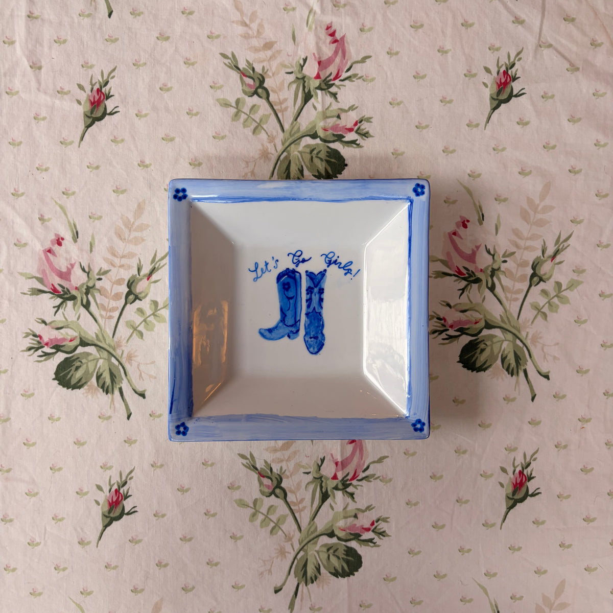 A square white ceramic dish with a blue rim and blue hand-painted calligraphy, placed on a pink floral tablecloth.