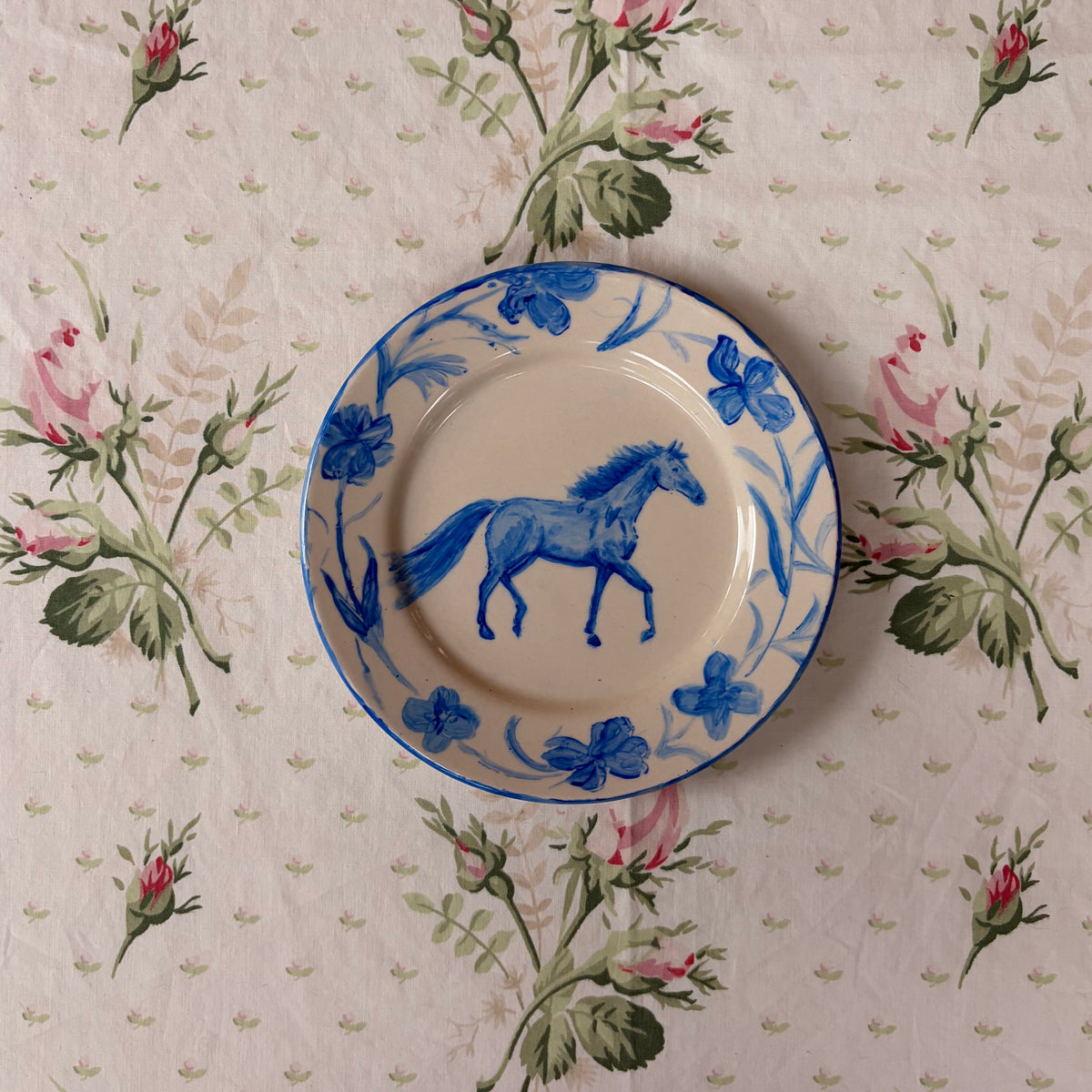 A round ceramic plate with a blue horse and floral border, placed on a beige tablecloth with a pink rose pattern.