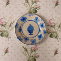 A round ceramic plate with a blue floral pattern and a central blue urn design, placed on a beige tablecloth with a pink rose print.