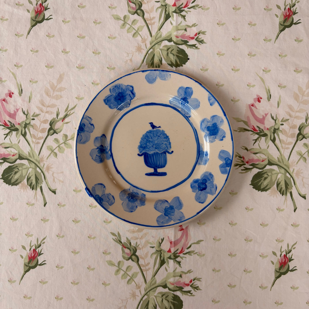 A round ceramic plate with a blue floral pattern and a central blue urn design, placed on a beige tablecloth with a pink rose print.