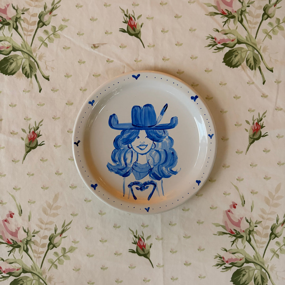 hand painted illustration in blue of a happy country singer cowgirl with a feather in her hat, her favorite bandana, bustier top, and a big smile. Framed in a dainty heart motif border on a small white plate. 