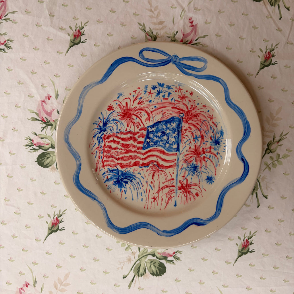 large hand painted fourth of July patriotic plate with fireworks behind a waving American flag, framed in a hand painted delicate blue bow ribbon border