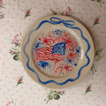 large hand painted fourth of July patriotic plate with fireworks behind a waving American flag, framed in a hand painted delicate blue bow ribbon border