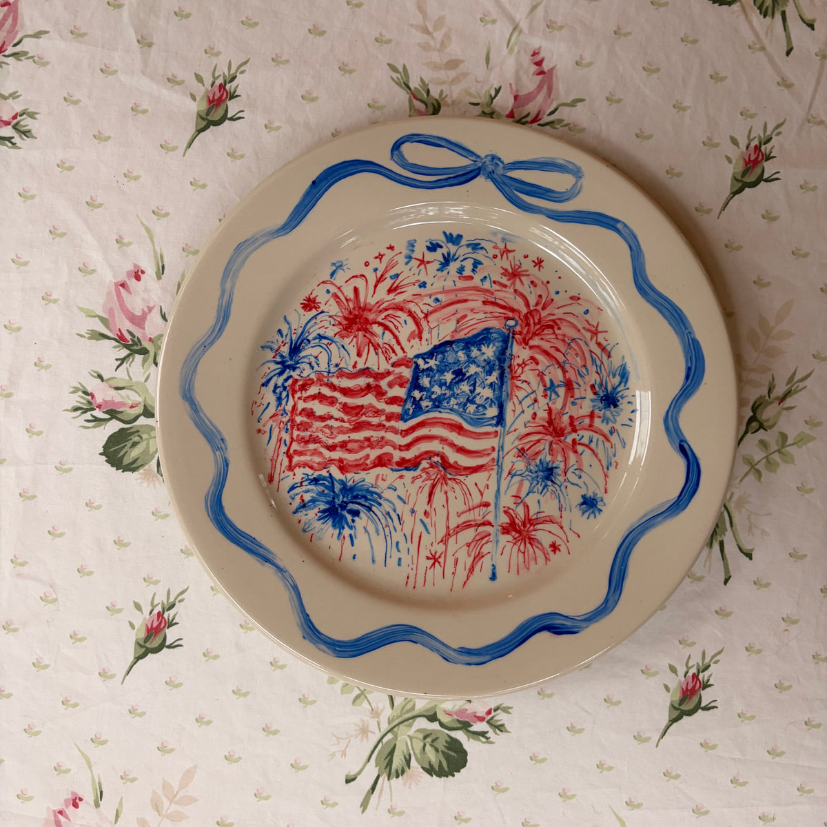 large hand painted fourth of July patriotic plate with fireworks behind a waving American flag, framed in a hand painted delicate blue bow ribbon border