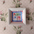 a green and brown hand painted palm tree with patriotic red and blue fireworks framed in red and blue scalloped border on a small square dish