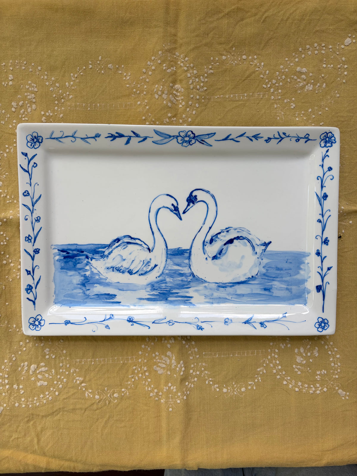 A rectangular white ceramic serving tray with a blue floral border and a central illustration of two swans facing each other in blue water, placed on a mustard yellow patterned fabric background.
