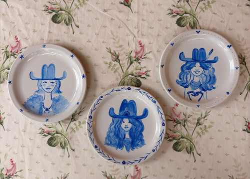 Three small white ceramic plates with blue hand-painted illustrations of women in cowboy hats, arranged on a beige floral tablecloth.